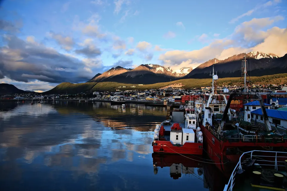 Barcos pesqueros rojos en el puerto de Ushuaia, con la ciudad y las montañas reflejadas en la bahía con luz dorada