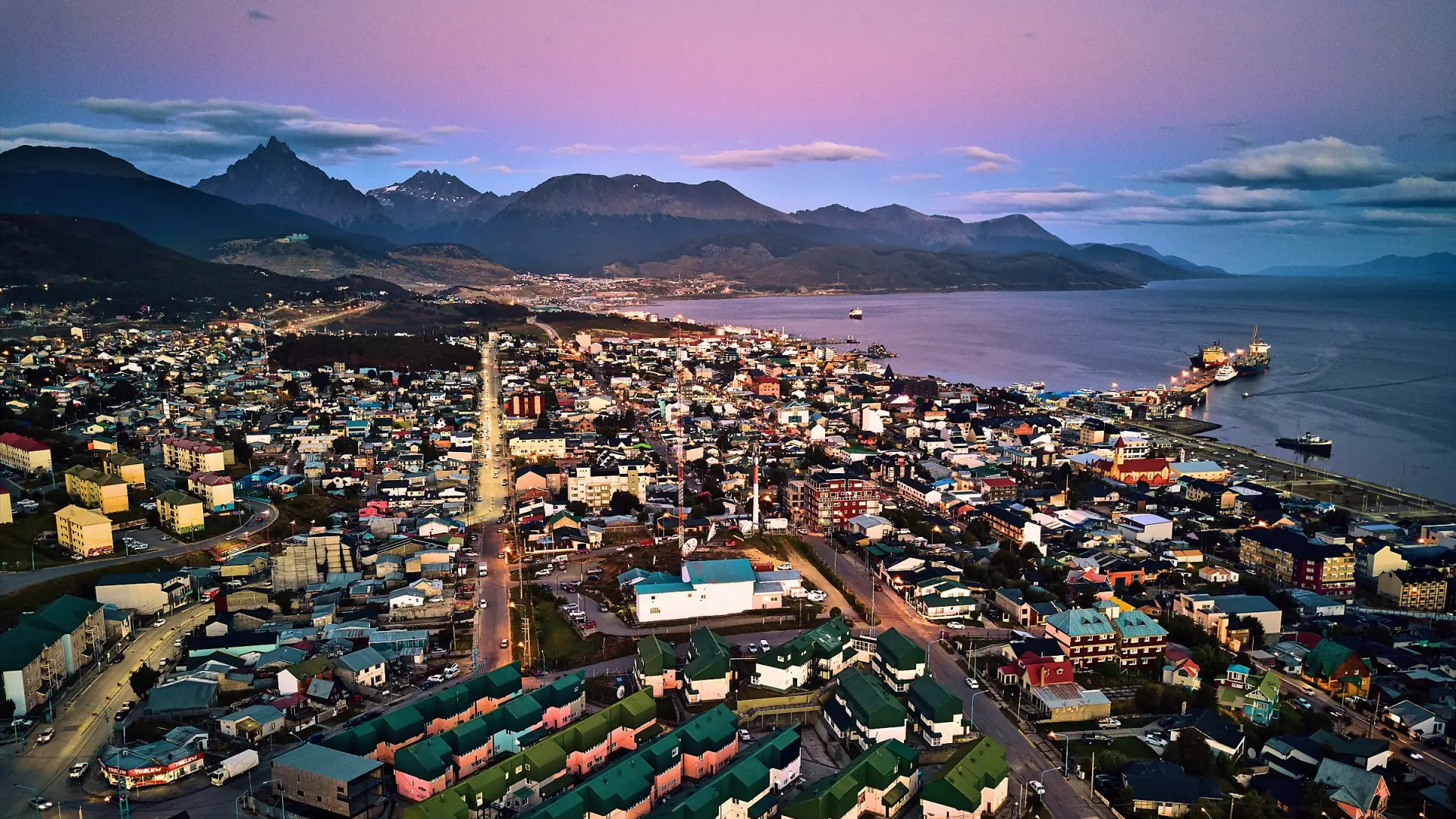 Ushuaia al anochecer vista desde el aire: la ciudad iluminada, el puerto sobre la bahía y las montañas bajo un cielo rosado