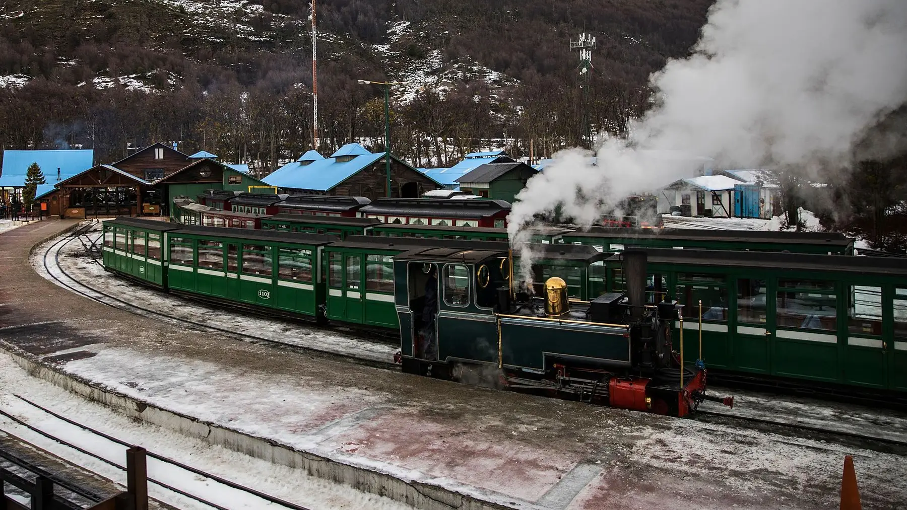 El tren a vapor en la estación, con vagones verdes, nieve y cerros en invierno
