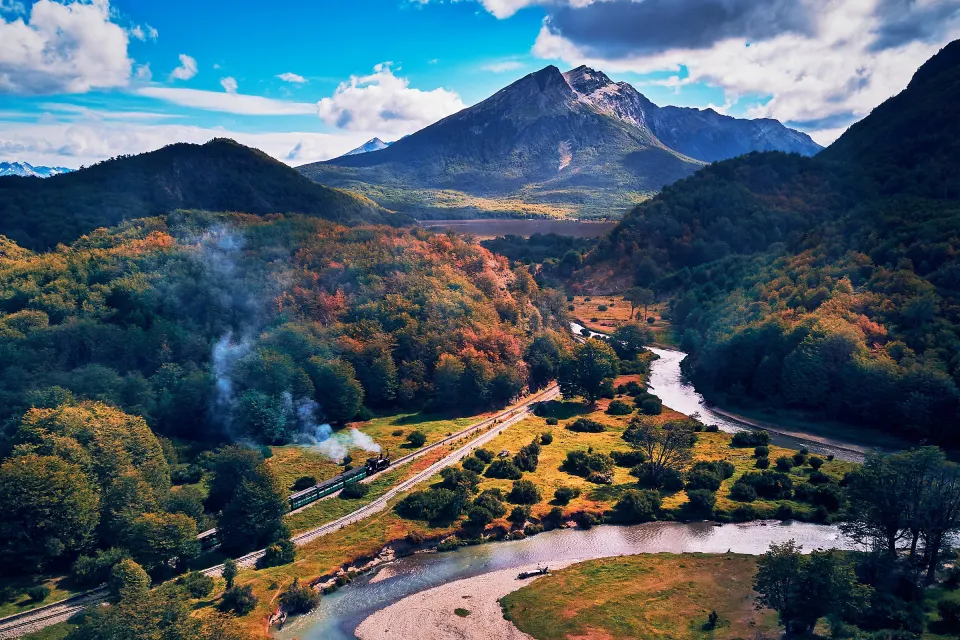 El Tren del Fin del Mundo con su columna de vapor, entre bosques de otoño y un río, en el Parque Nacional Tierra del Fuego