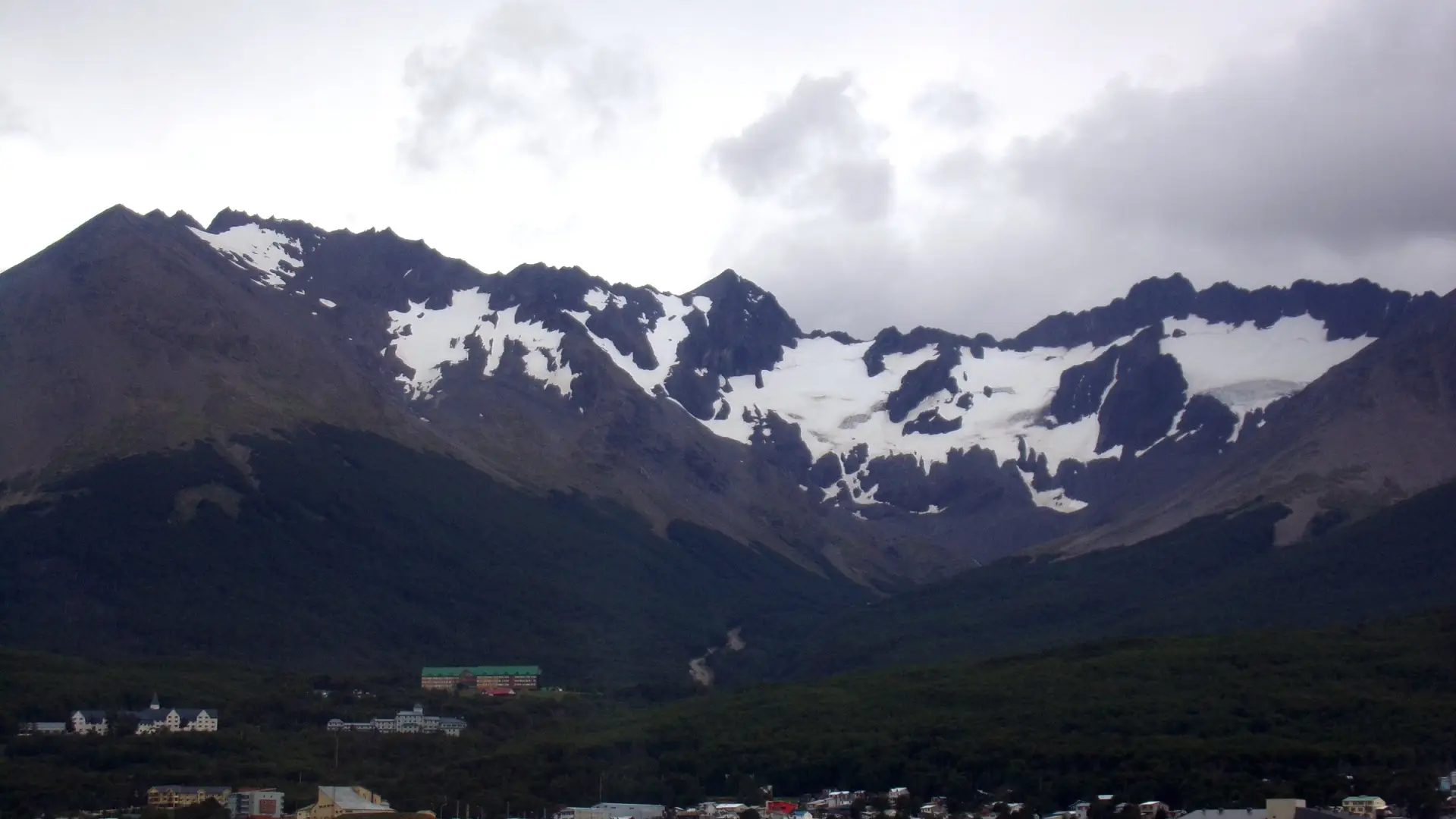El circo del Glaciar Martial con nieve entre cumbres oscuras, sobre el bosque y las casas de Ushuaia en un día nublado