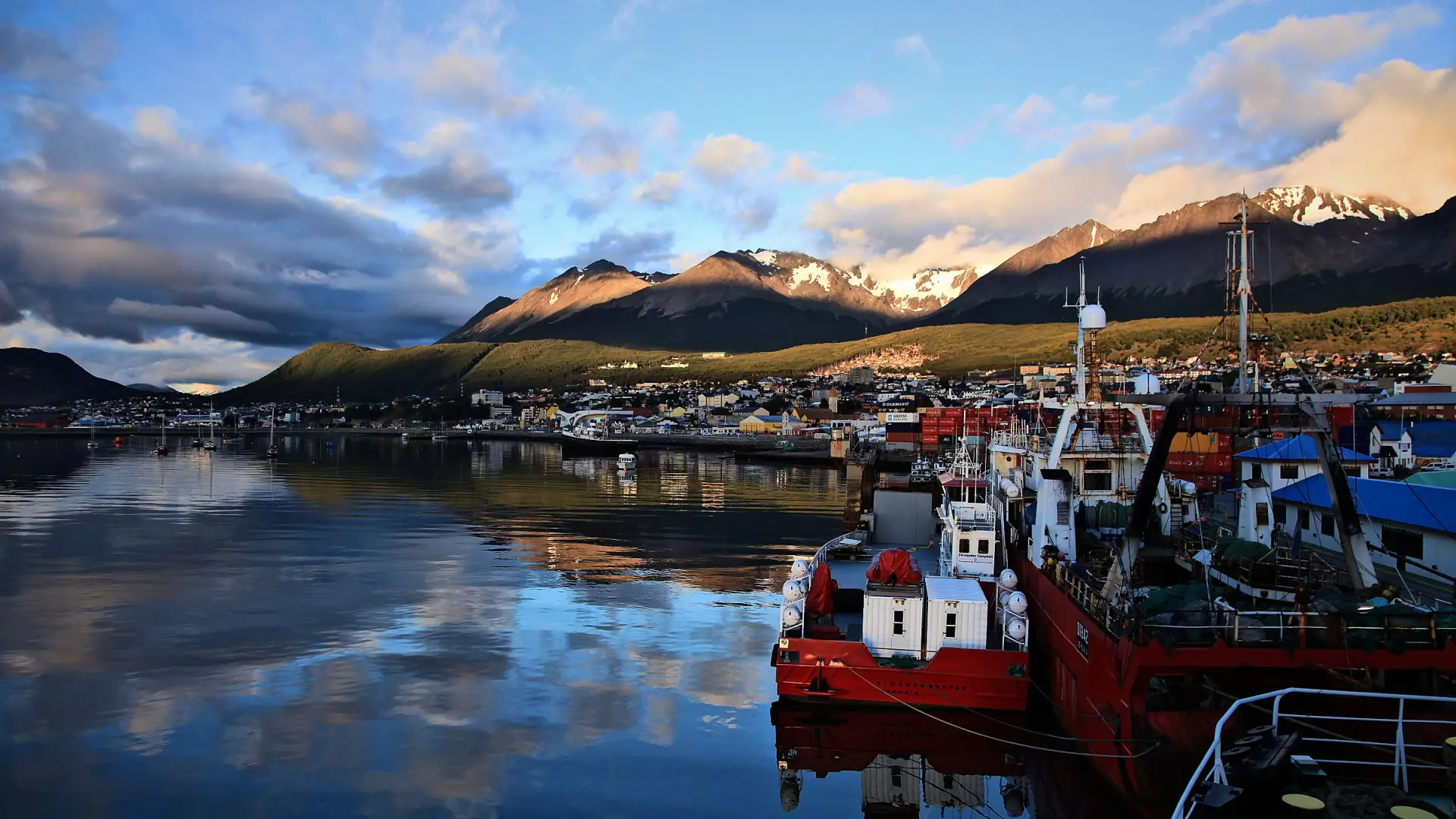 Barcos pesqueros rojos en el puerto de Ushuaia, con la ciudad y las montañas reflejadas en la bahía con luz dorada