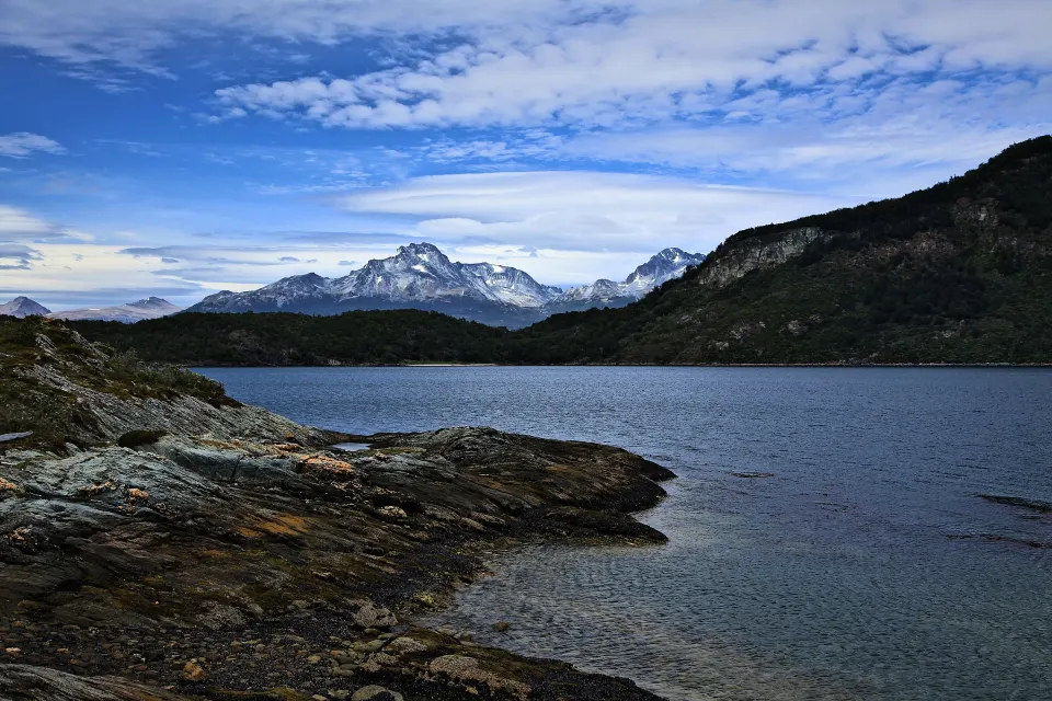 La costa de rocas de la Bahía Lapataia, en el Parque Nacional Tierra del Fuego, con bosque y montañas nevadas