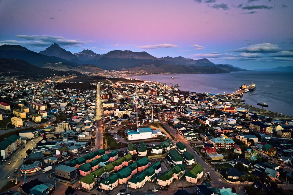 Ushuaia al anochecer vista desde el aire: la ciudad iluminada, el puerto sobre la bahía y las montañas bajo un cielo rosado