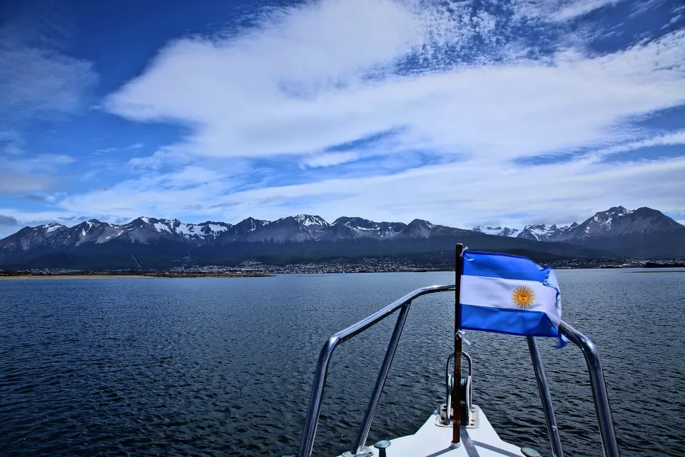 La proa de un barco con la bandera argentina navegando el Canal Beagle, con Ushuaia y las montañas nevadas al fondo