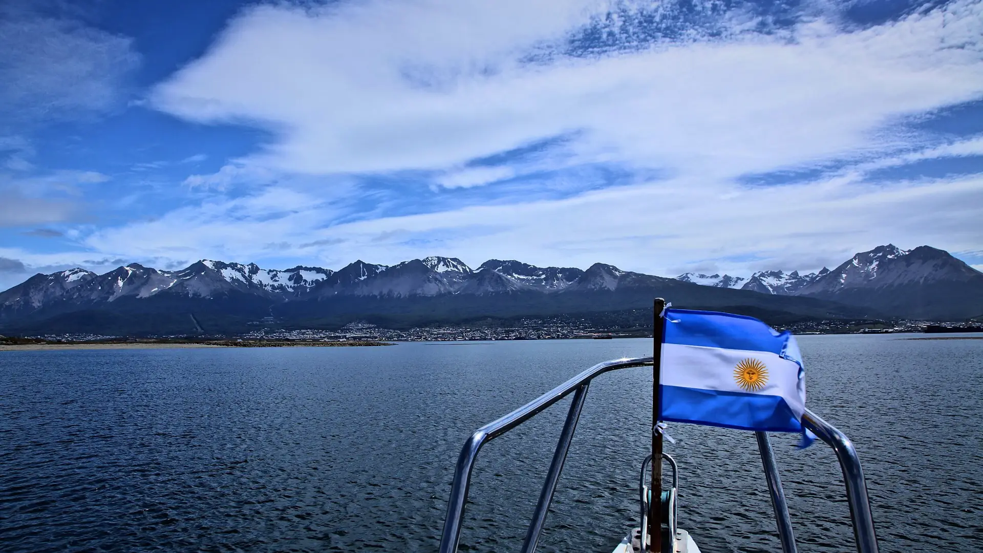 La proa de un barco con la bandera argentina navegando el Canal Beagle, con Ushuaia y las montañas nevadas al fondo